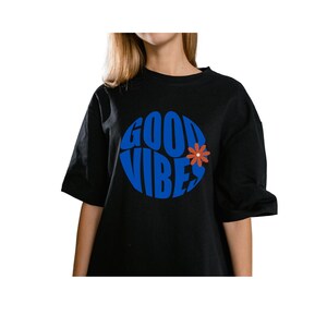 Könnte beinhalten: Schwarzes T-Shirt mit einem blauen, kreisförmigen Aufdruck mit der Aufschrift "GOOD VIBES" und einer kleinen orangefarbenen Blume. Das Shirt hat kurze Ärmel und einen Rundhalsausschnitt.
