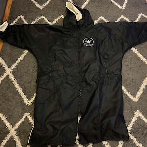 Puede incluir: Bata de cambio negra con capucha y mangas largas, colocada sobre una alfombra gris estampada. La bata tiene una cremallera completa, ribetes blancos y un forro de forro polar blanco en la capucha y las mangas. Un logotipo circular con texto está en el pecho.