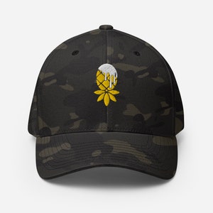 Puede incluir: Una gorra de béisbol de camuflaje negro y verde con una piña amarilla y glaseado blanco bordado en la parte delantera.