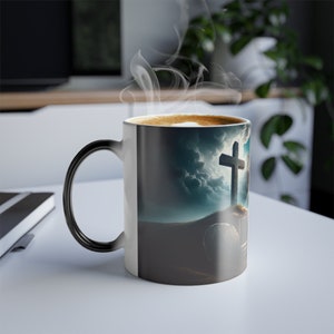 May include: A black and white heat-sensitive mug with a cross and a stone tomb on a cloudy background. The mug is filled with coffee and steam is rising from the top.