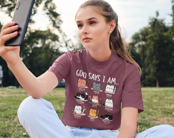 May include: A woman wearing a maroon t-shirt with a graphic of cats and the text "God Says I Am..."  The graphic features a list of positive affirmations about a person's identity, each represented by a different cat.