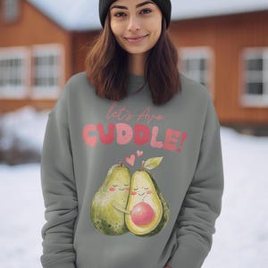 Può includere: Una felpa grigia con una grafica di due avocado che si abbracciano. Il testo sulla felpa dice "Let's Avo Cuddle!" con lettere rosa e rosse.