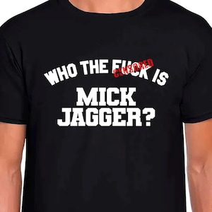 Puede incluir: Camiseta negra con la frase "WHO THE F*CK IS MICK JAGGER?" en letras blancas. La palabra "F*CK" está parcialmente cubierta con un sello rojo. Cuello redondo clásico.