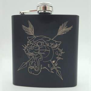 Puede incluir: Frasco de metal negro con un diseño grabado en oro de la cabeza de una pantera con dos flechas cruzándose detrás de ella.