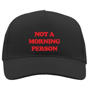 Puede incluir: Gorra de béisbol negra con el texto rojo "NOT A MORNING PERSON". La gorra tiene una visera curva y una corona estructurada. Un accesorio casual para el uso diario.