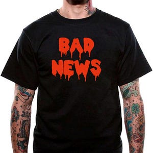 以下が含まれることがあります： オレンジ色の滴る文字で「BAD NEWS」と書かれた黒いTシャツ。半袖シャツは柔らかい素材で作られています。デザインは胸の中央に配置されています。