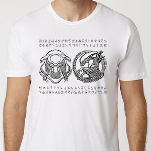 May include: White t-shirt featuring black graphic designs of a Predator and an Alien, with text in an unknown language. The shirt has a crew neck and short sleeves. The design includes circular and linear elements.