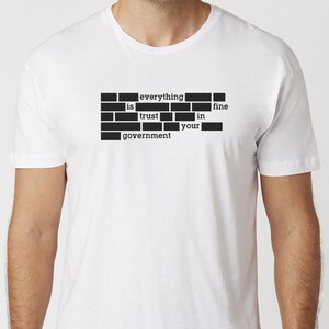 Può includere: T-shirt bianca con un design grafico nero. Il design è un blocco rettangolare con le parole "everything is fine trust in your government" in un carattere di macchina da scrivere.