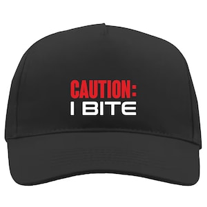 Puede incluir: Gorra de béisbol negra con el texto "CAUTION: I BITE" en rojo y blanco. La gorra tiene una visera curva y una corona estructurada. El texto está centrado en la parte delantera.