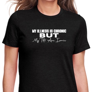 Puede incluir: Una camiseta negra de manga corta con texto blanco que dice MY ILLNESS IS CHRONIC BUT My Tits Are Iconic. La prenda tiene un cuello redondo y un corte estándar, con letras en bloque en la línea superior y una fuente cursiva debajo.