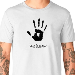 May include: White t-shirt with a black handprint and the text "We Know?" printed on the front.