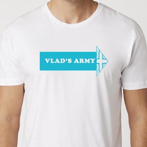 Może przedstawiać: Biała koszulka z turkusowym wzorem strzałki. Strzałka zawiera napis "VLAD'S ARMY" białymi literami. Grot strzałki ma wzór krzyża. Koszulka jest wykonana z miękkiego materiału.