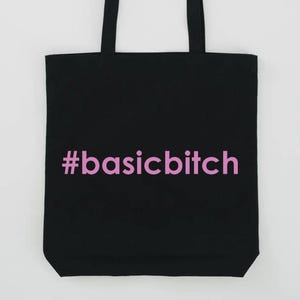 Puede incluir: Bolso tote negro con el texto rosa "#basicbitch". El bolso tiene dos asas negras y está hecho de un material resistente. El texto está centrado en la parte delantera del bolso.