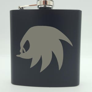 Puede incluir: Frasco de metal negro con una tapa plateada y una silueta gris de la cabeza de Sonic the Hedgehog con púas.