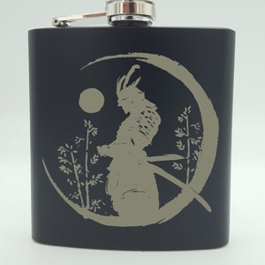 May include: Black stainless steel flask with a silver cap and a design of a samurai warrior with a sword in a circle with a moon and trees.