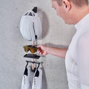 May include: A white bicycle helmet, a pair of gold-rimmed sunglasses, and two white cycling shoes are hanging on a black wall-mounted rack.