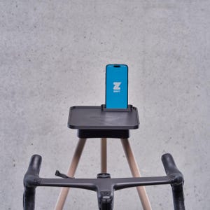 Könnte beinhalten: Ein schwarzer Telefonständer mit einem Holz-Dreibeinstativ. Der Telefonständer hat ein Tablett für ein Telefon und einen Schlitz für Ladekabel. Ein schwarzer Fahrradlenker befindet sich im Vordergrund. Der Telefonbildschirm zeigt die Zwift-App an.