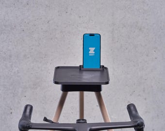 May include: A black phone stand with a wooden tripod base. The phone stand has a tray for a phone and a slot for charging cables. A black bicycle handlebar is in the foreground. The phone screen displays the Zwift app.