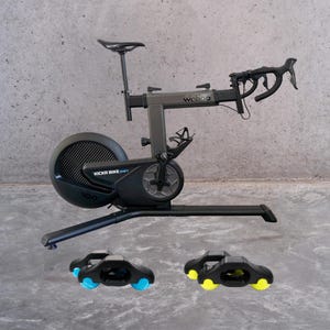 Könnte beinhalten: Ein schwarzes Indoor-Fahrrad mit einem Wahoo-Logo am Rahmen. Das Fahrrad hat ein schwarzes Schwungrad mit dem Text "KICKR BIKE SHIFT 100" darauf. Das Fahrrad steht auf einem grauen Betonboden mit zwei schwarzen und blauen Radblöcken.
