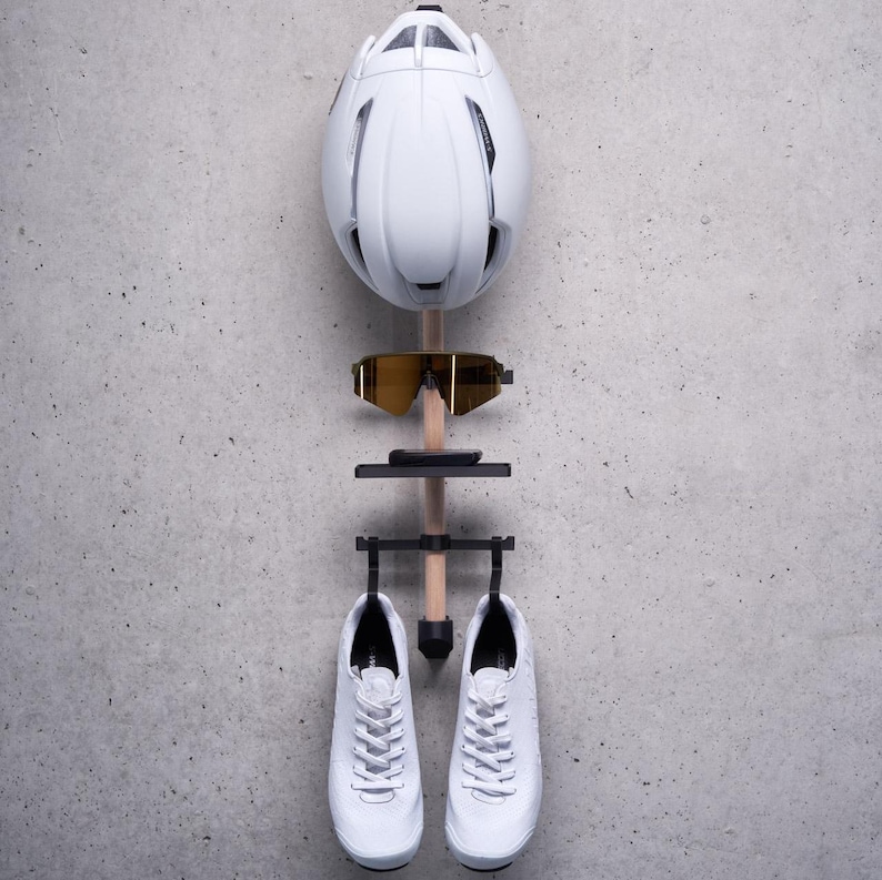 May include: A white bicycle helmet, a pair of yellow sunglasses, a black phone holder, and two white cycling shoes are hanging on a wooden rack against a gray background.