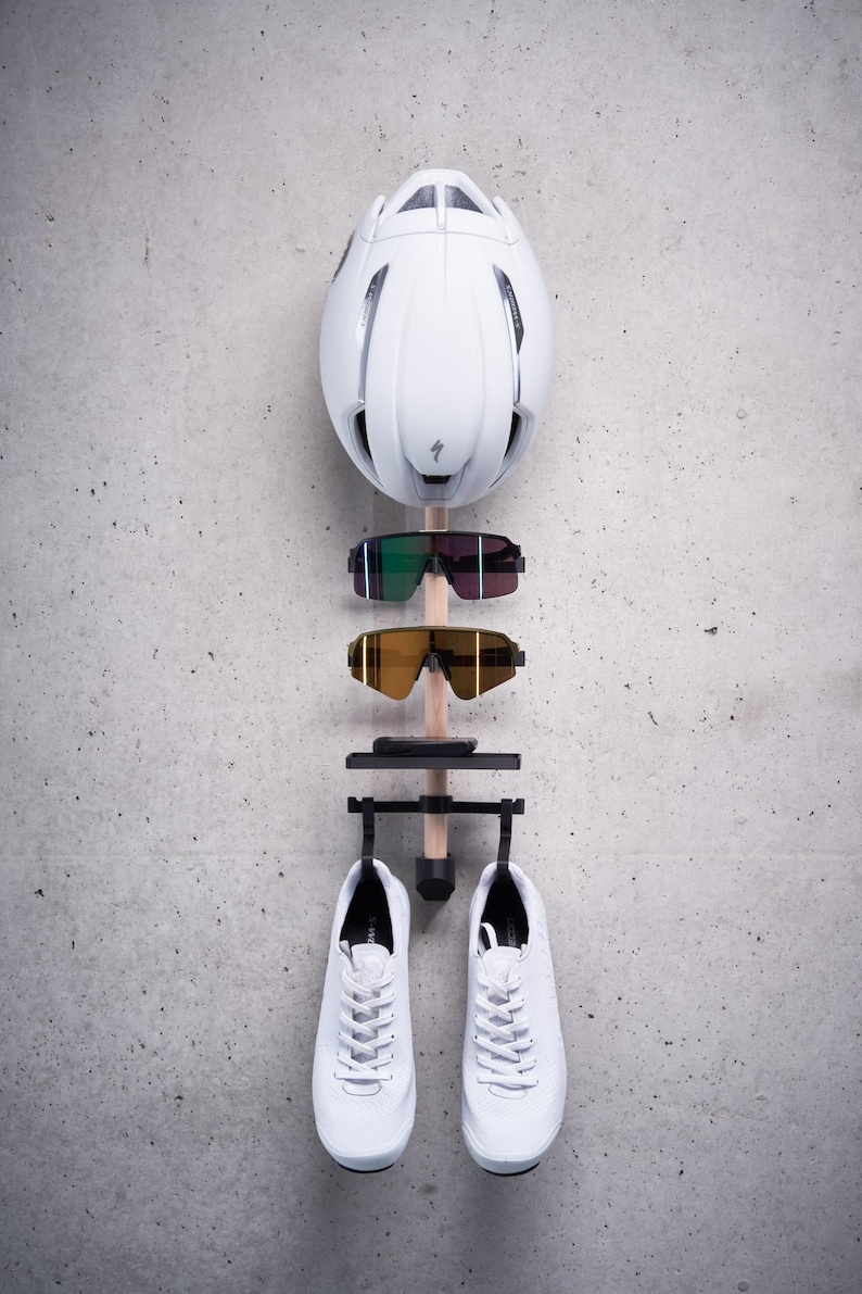 May include: White cycling helmet, two pairs of sunglasses, and a pair of white sneakers hanging on a wooden rack against a gray concrete wall.