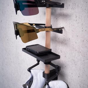 May include: A black wall-mounted organizer with a wooden dowel, holding two pairs of white sneakers, a black phone case, and two pairs of sunglasses. The sunglasses are both black framed with one pair having blue lenses and the other pair having gold lenses. The sunglasses are labeled "Oakley".