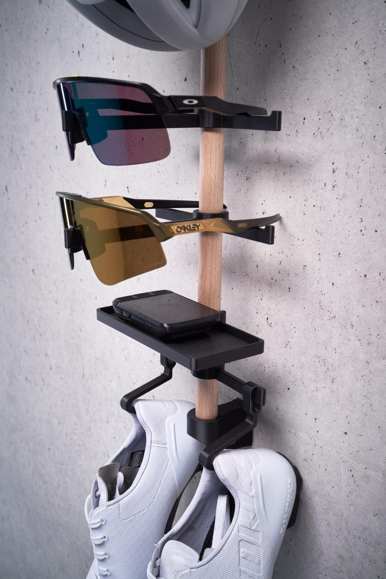 May include: A black wall-mounted organizer with a wooden dowel, holding two pairs of white sneakers, a black phone case, and two pairs of sunglasses. The sunglasses are both black framed with one pair having blue lenses and the other pair having gold lenses. The sunglasses are labeled "Oakley".