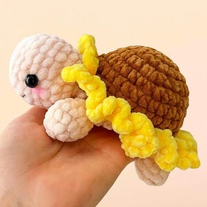 Puede incluir: Una tortuga de ganchillo con un diseño de girasol. La tortuga tiene un caparazón marrón claro, una cabeza y patas de color crema y detalles de girasol amarillos. El texto "little sunflower turtle no sew - pdf pattern" está en la parte superior de la imagen.