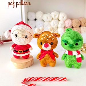 Puede incluir: Conjunto navideño de ganchillo con Papá Noel, un reno y el Grinch. La figura de Papá Noel es roja y blanca, el reno es marrón con un lazo rojo y el Grinch es verde con una bufanda a rayas. Un bastón de caramelo está en primer plano.