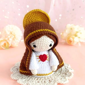 Puede incluir: Muñeca de ganchillo de la Virgen María, con un tocado marrón y dorado y un vestido blanco con un corazón rojo. La muñeca está sobre una base decorativa de ganchillo. El texto "VIRGEN DOLOROSA" está en la parte superior.