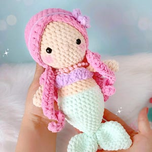 Puede incluir: Muñeca de sirena de ganchillo con pelo rosa y cola verde claro. La muñeca tiene un gorro rosa y un collar de perlas. El texto "Low-Sew Amigurumi Pattern" está en la parte superior de la imagen.