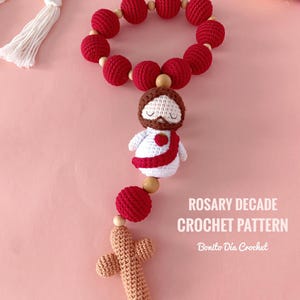 Puede incluir: Un rosario de ganchillo hecho a mano con cuentas rojas y una pequeña figura de Jesús. El rosario incluye una cruz de ganchillo y cuentas de madera. El texto "Rosary Decade Crochet Pattern" es visible.
