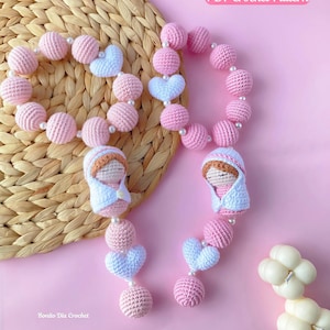May include: Two crochet rosary-style garlands featuring pink spheres, white heart-shaped beads, and small white pearls. Each garland includes a small crocheted figure wearing a white hood and brown hair. Text reads PDF Crochet Pattern.