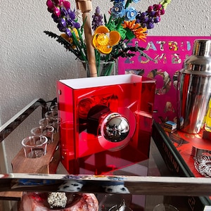 May include: A red acrylic bar cart with a chrome sphere detail. The cart holds a cocktail shaker, shot glasses, and a vase of colourful artificial flowers. A book with pink cover is also visible.