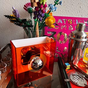 May include: An orange acrylic bar caddy with a mirrored sphere and a white interior. The caddy sits on a glass surface with cocktail tools, a cocktail shaker, and a bouquet of colourful flowers in the background.