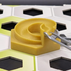 Könnte beinhalten: Eine gelbe Kunststoff-Murmelbahnkomponente mit einer gebogenen Bahn und einer quadratischen Basis. Silberne Kunststoffzangen sind in der Nähe der Bahn positioniert. Die Komponente befindet sich auf einer sechseckigen Fliesenoberfläche mit weißen, grauen und grünen Akzenten. Der Hintergrund ist schwarz.