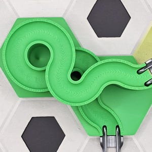 Może przedstawiać: Zielony tor dla kulek ze spiralnymi i zakrzywionymi sekcjami, połączony ze srebrnym mechanizmem startowym. Tor znajduje się na białej powierzchni z czarnymi i żółtymi sześciokątnymi wzorami.