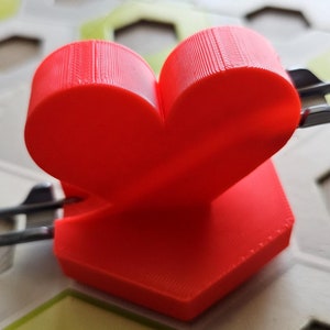 Könnte beinhalten: Ein rotes herzförmiges Objekt mit einer sechseckigen Basis, das auf einer weißen und grünen gemusterten Oberfläche liegt. Das Herz ist aus einem 3D-gedruckten Material gefertigt und hat eine strukturierte Oberfläche.