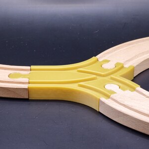 Puede incluir: Vía de tren de juguete de madera con un conector en forma de Y de plástico amarillo. La vía es de color marrón claro, con un diseño curvo. El conector permite que la vía se divida en dos direcciones.