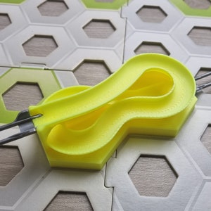 Könnte beinhalten: Ein gelbes Kunststoff-Streckenstück für ein Murmelbahn-Spiel. Die Strecke ist in Form einer Acht geformt und hat eine strukturierte Oberfläche. Die Strecke ist an einer grauen Metallschiene befestigt.