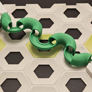 Puede incluir: Una pieza de pista verde y curvada está conectada a dos formas geométricas blancas sobre una base de baldosas hexagonales. La base tiene baldosas blancas y amarillas con recortes hexagonales negros. Parece un juego o un rompecabezas.