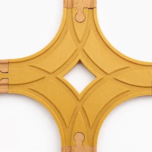 Pode incluir: Uma peça de intersecção de trilhos de trem de brinquedo de madeira com um conector de plástico amarelo mostarda. A pista é castanho-claro e o conector tem um design geométrico. As peças encaixam-se para criar uma ferrovia.