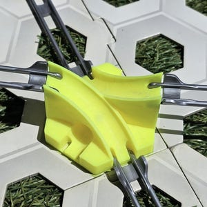 Könnte beinhalten: Nahaufnahme einer gelben Kunststoffkomponente mit gebogenen Kanälen, verbunden durch silberne Metallstäbe. Sie liegt auf sechseckigen Fliesen mit Graseinsätzen, möglicherweise für die Landschaftsgestaltung.