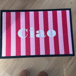 May include: A rectangular doormat with a black border features alternating vertical stripes of light and dark pink. The word "Ciao" is printed in white across the center of the mat. The mat is designed for indoor or outdoor use.
