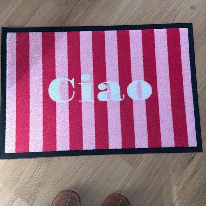 May include: A rectangular doormat with a black border features alternating vertical stripes of light and dark pink. The word "Ciao" is printed in white across the center of the mat. The mat is designed for indoor or outdoor use.