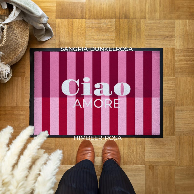 May include: A rectangular doormat with a black border, featuring alternating vertical stripes of light and dark pink. The words "Ciao AMORE" are in white, centered on the mat. The text "SANGRI-DUNKELROSA" and "HIMBEER-ROSA" are also visible.