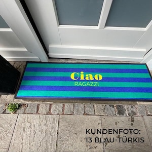 May include: A rectangular doormat with horizontal stripes in shades of blue and green. The words "Ciao RAGAZZI" are in yellow, centered on the mat. The mat is placed in front of a white door.