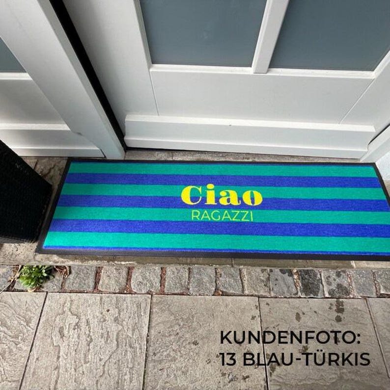 May include: A rectangular doormat with horizontal stripes in shades of blue and green. The words "Ciao RAGAZZI" are in yellow, centered on the mat. The mat is placed in front of a white door.