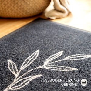 May include: A dark gray doormat with a white leaf design. The mat has a textured surface and the text "FUSSBODENHEIZUNG GEEIGNET" is printed on the bottom right corner. The mat is suitable for floor heating.
