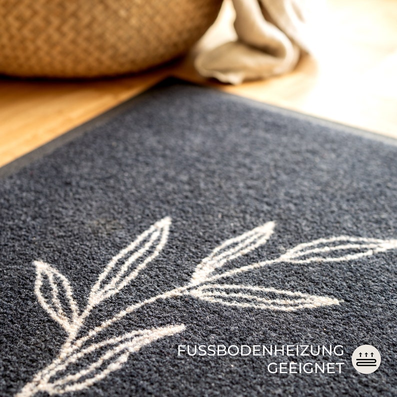 May include: A dark gray doormat with a white leaf design. The mat has a textured surface and the text "FUSSBODENHEIZUNG GEEIGNET" is printed on the bottom right corner. The mat is suitable for floor heating.